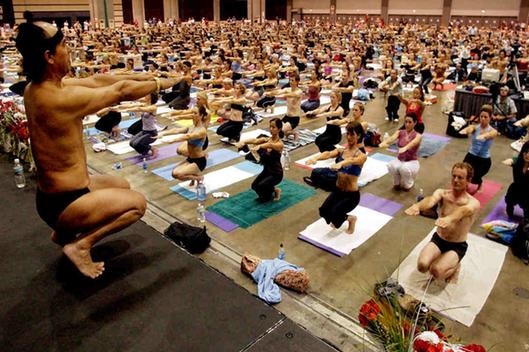 Man with a headband on teaches a large room of people yoga.