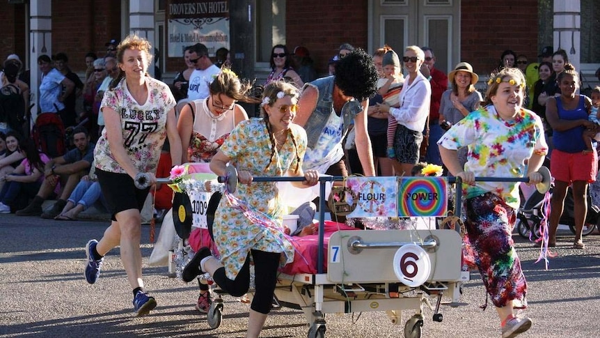 Participants in hippy-style fancy dress push a decorated hospital bed through the main street of Moora.