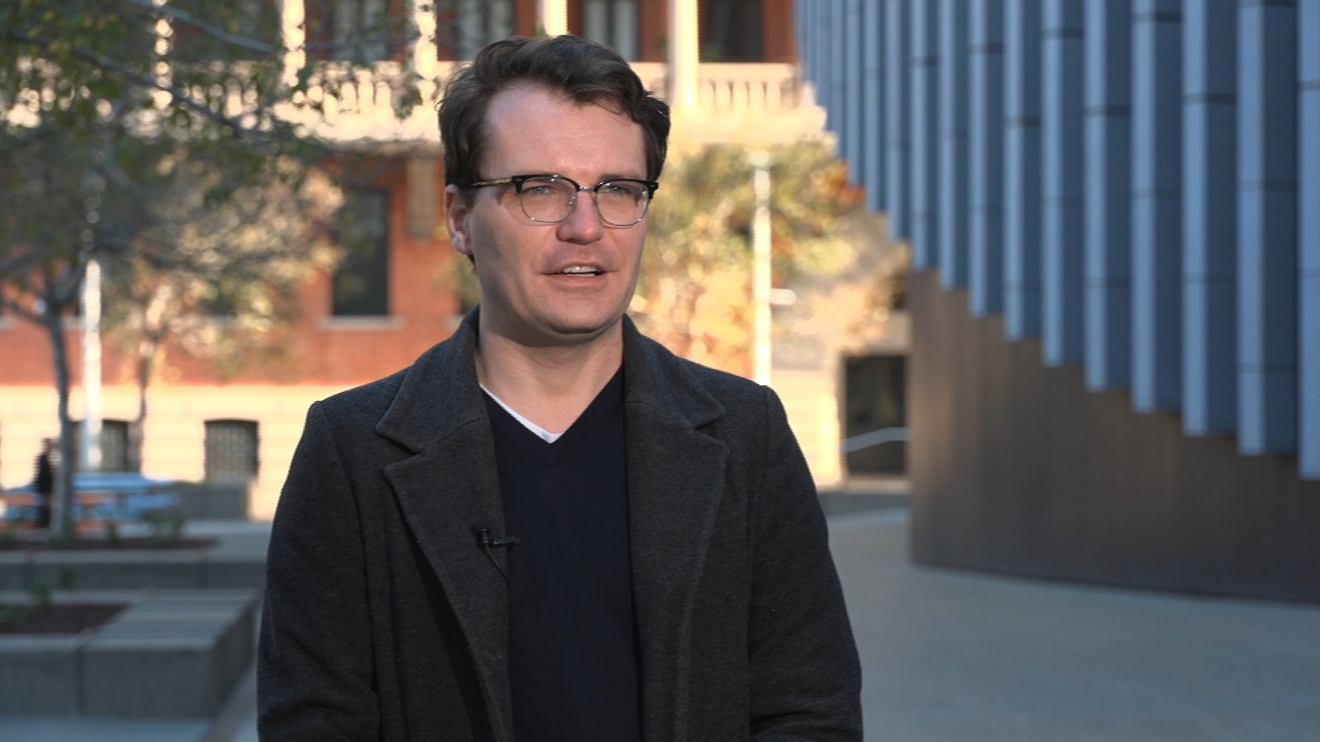 A man with black frame glasses and slicked back hair dressed in black clothing.
