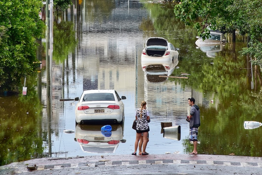 Two people stand in front of a flooded road in Herston, with two cars partially submerged.