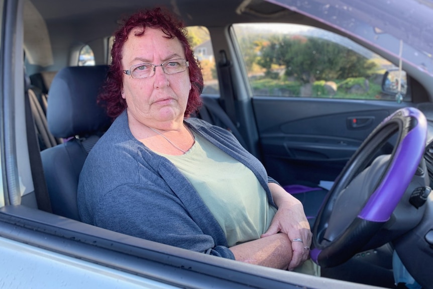 A middle-aged woman with red hair sits behind the wheel of a car