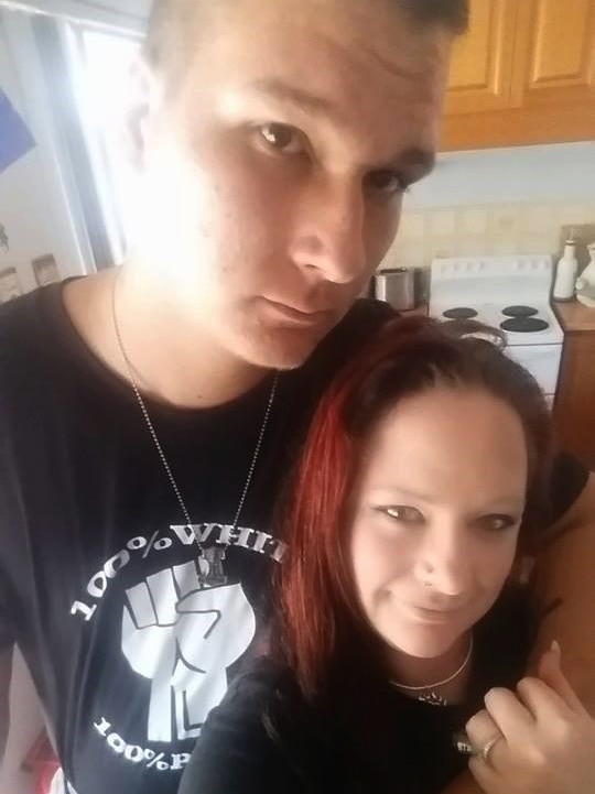 A man and a woman stand in a kitchen with their arms around one another, with the man wearing a 'white power' t-shirt.