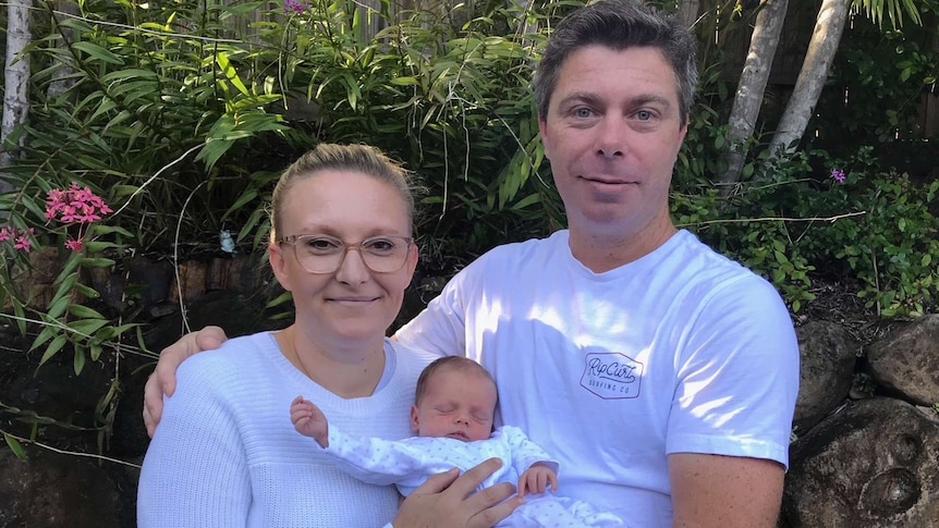 Man and woman holding a newborn baby in front of a shrub