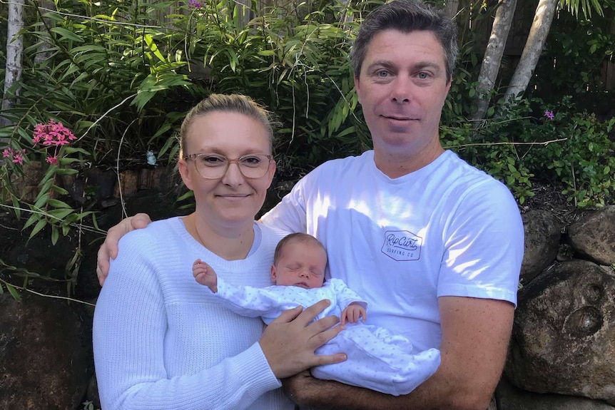 Man and woman holding a newborn baby in front of a shrub