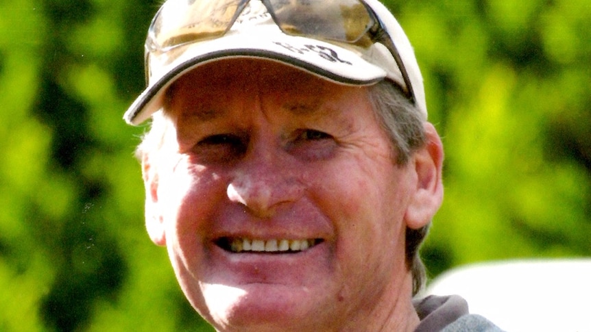 A man wearing a grey top smiles happily at the camera, in front of green trees.