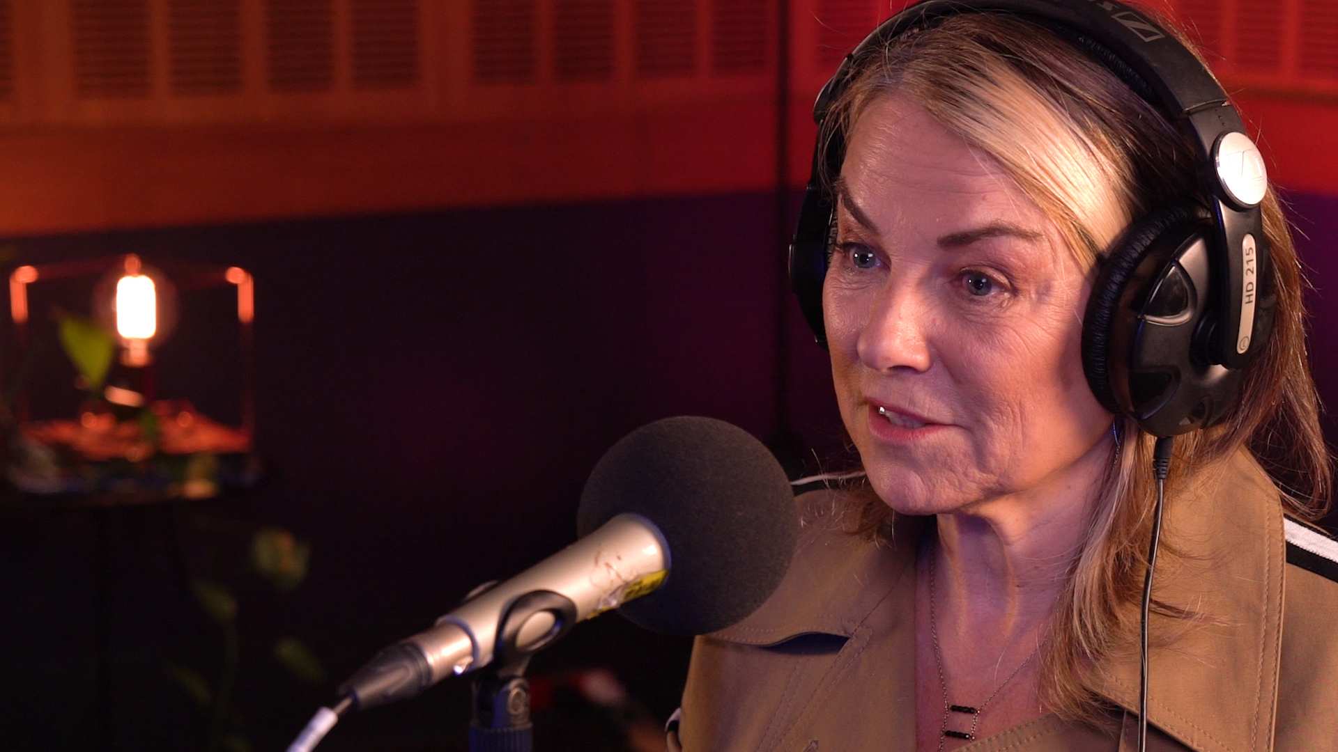 A woman with blonde hair and a beige jacket wearing headphones and speaking into a microphone.