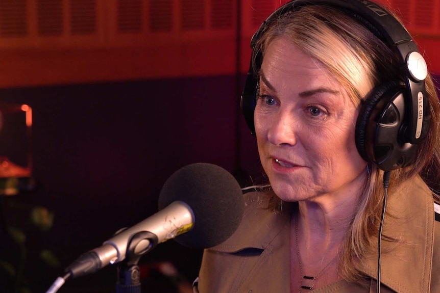 A woman with blonde hair and a beige jacket wearing headphones and speaking into a microphone.