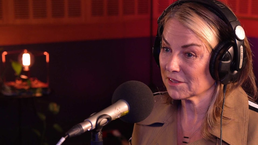 A woman with blonde hair and a beige jacket wearing headphones and speaking into a microphone.