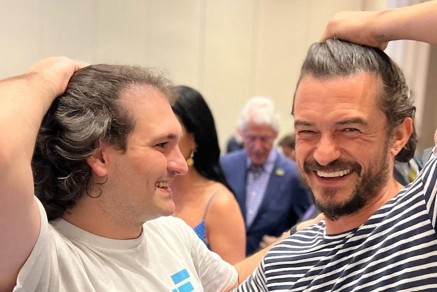 Sam Bankman-Fried and Orlando Bloom push their hair back and laugh 