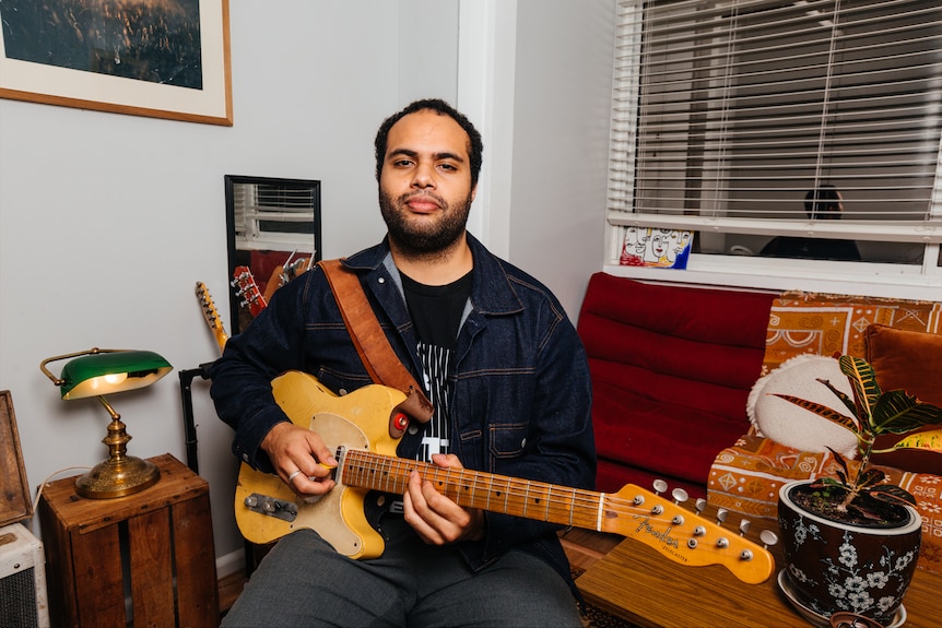 Joji sits on a chair holding a yellow electric guitar