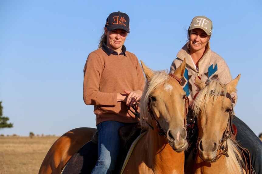Two women wearing trucker hats smile sitting on horseback