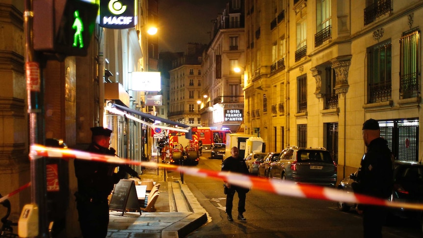 Police tape streches across a street in France where police officers with guns stand.