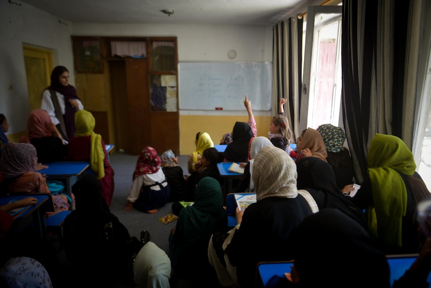 From the back of the classroom, Sodaba can be seen standing at the front of the class while two children put hands up.