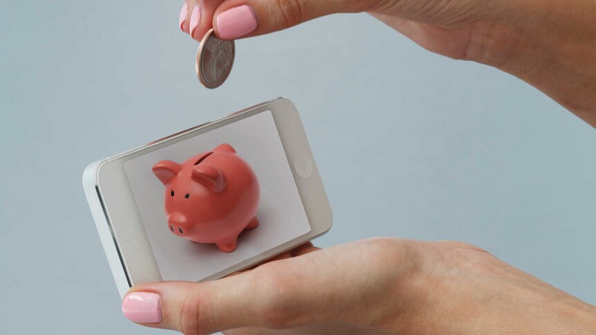 A woman holds a coin above a phone with an image of a piggy bank on it.