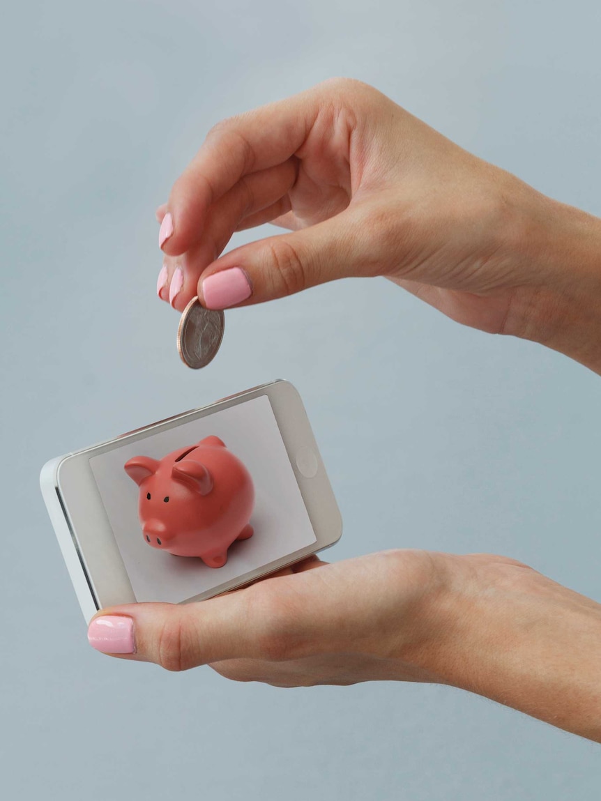 A woman holds a coin above a phone with an image of a piggy bank on it.