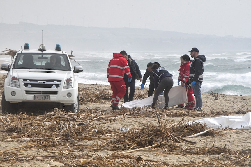Wood lies on the beach as a car and emergency workers cover a body in a white sheet.