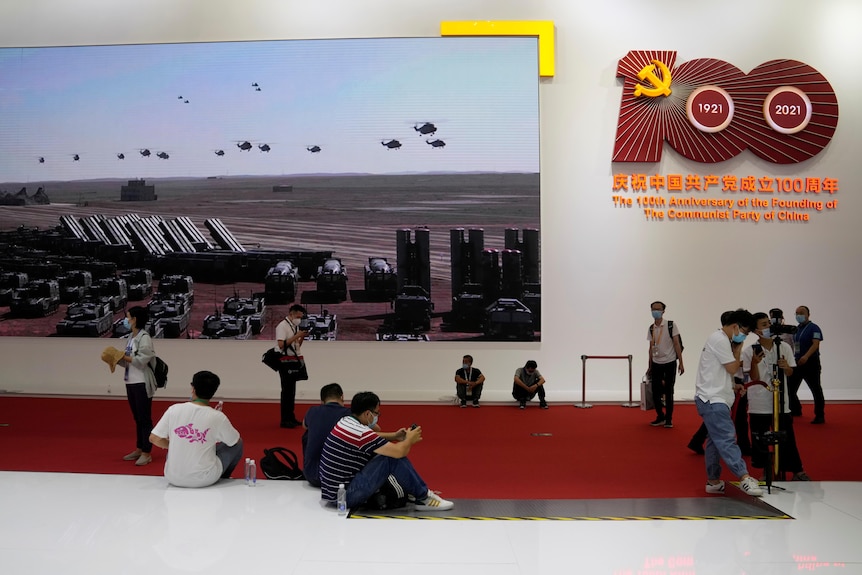 People sit on the ground and stand around in front of a screen depicting Chinese military hardware