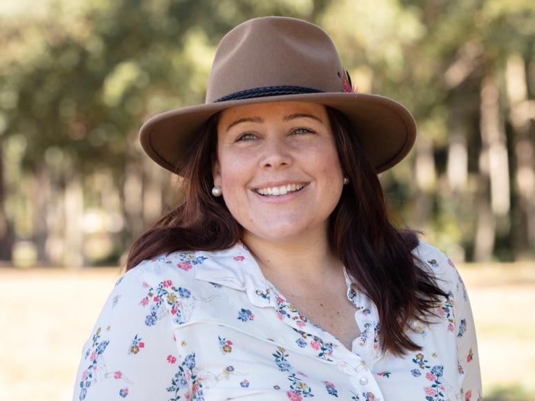 A young woman in an akubra is smiling, the background is blurred but she's standing in front of gum trees