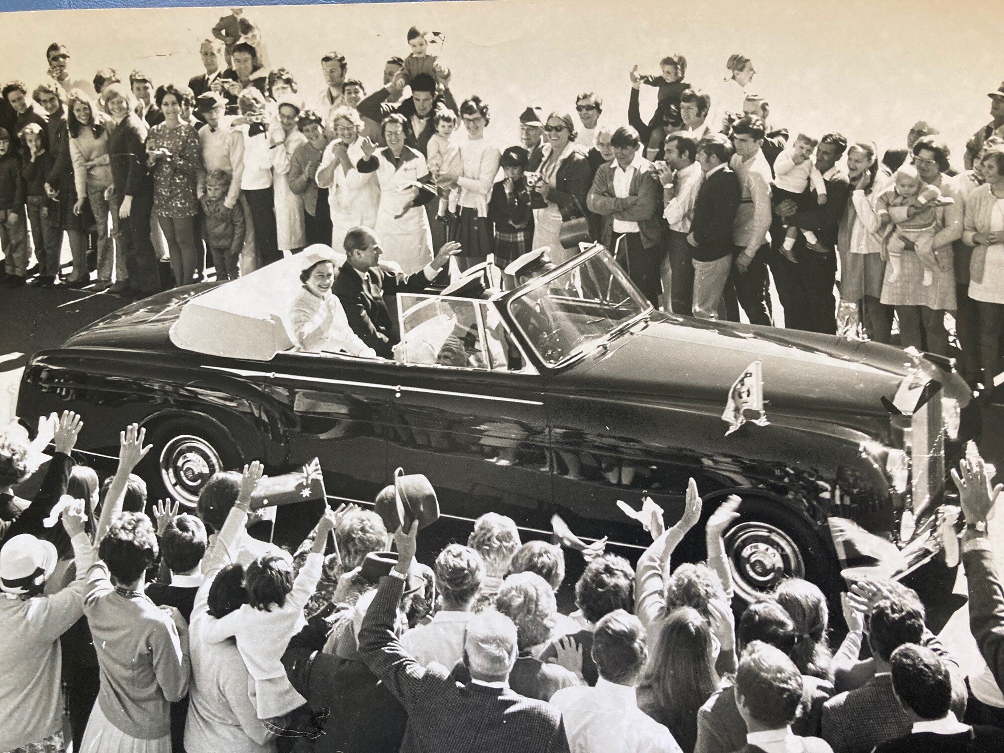 a black and white picture of the queen sitting in a car and waving as she arrives in a town surrounded by locals