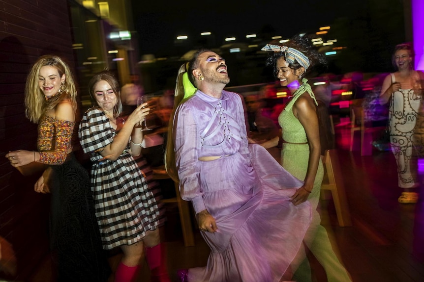 A group of people in vibrant and expressive clothing laugh as they dance at a gallery event.