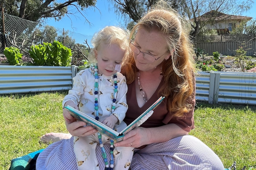 A mother and toddler read a book in the sunshine in a backyard