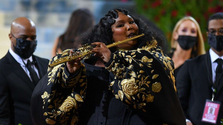 A woman in a large black gown with golden leaf and rose embroidery plays a golden flute decorated with golden leaves.