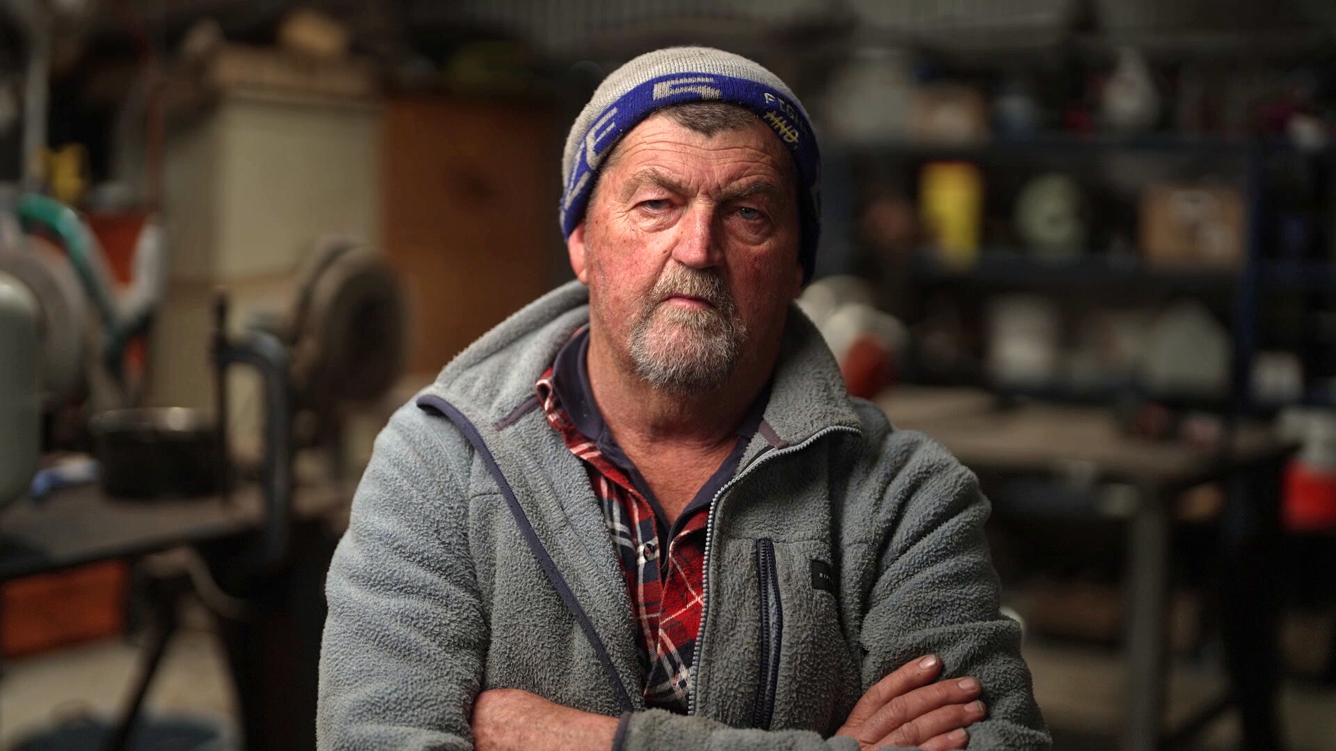 An older man with a beanie, grey beard and grey polar fleece zip up jumper sits in a workshop with his arms crossed.