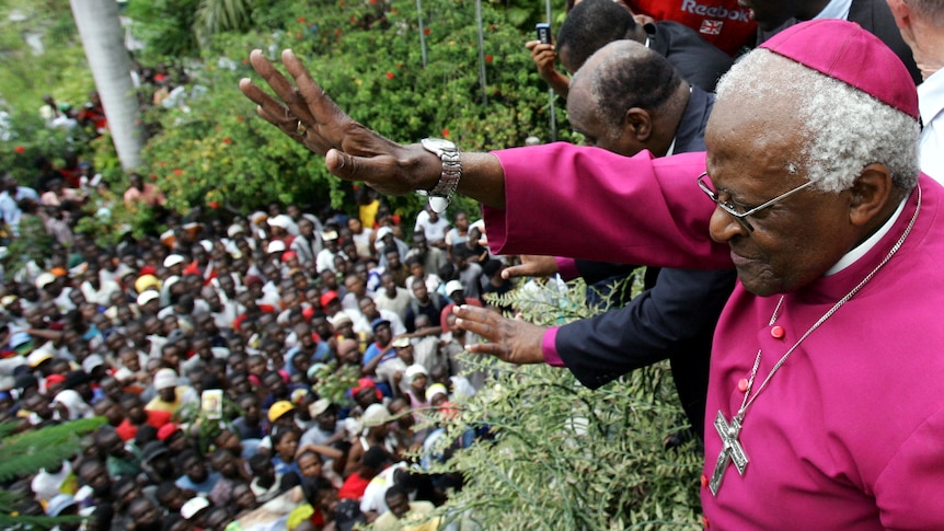 Archbishop Desmond Tutu waves to crowd of black people from elevated position, wearing red dress