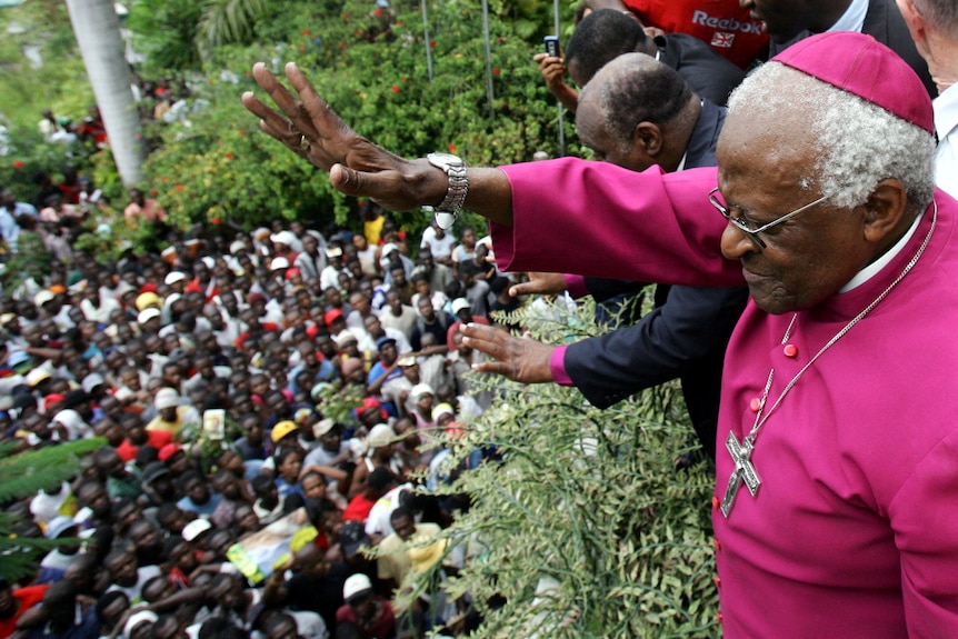 Archbishop Desmond Tutu waves to crowd of black people from elevated position, wearing red dress