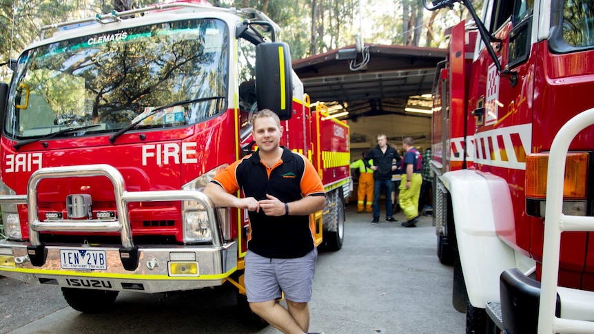 A man stands between two fire trucks outside a shed surrounded by trees.