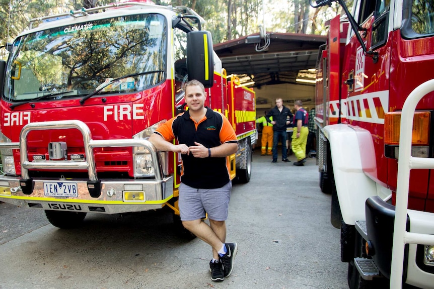 A man stands between two fire trucks outside a shed surrounded by trees.