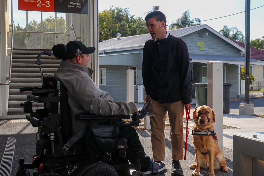 a man in a wheelchair and a man with a guide dog speaking to each other on a train platform