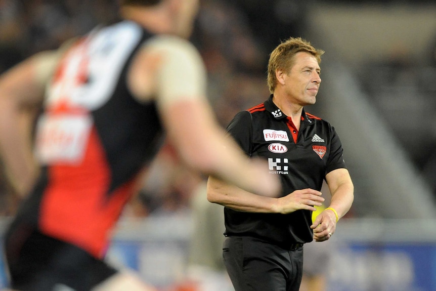 A blurred footballer runs through the frame in front of Mark Thompson on the MCG.