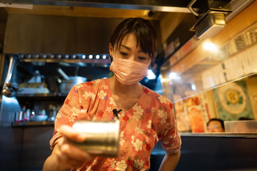 A young Japanese woman in a face mask seasoning meat with a salt shaker 