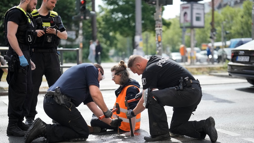 Police with hammers and chisels work to free the hand of an activist sitting with hi-vis orange vest with hand glued to road.