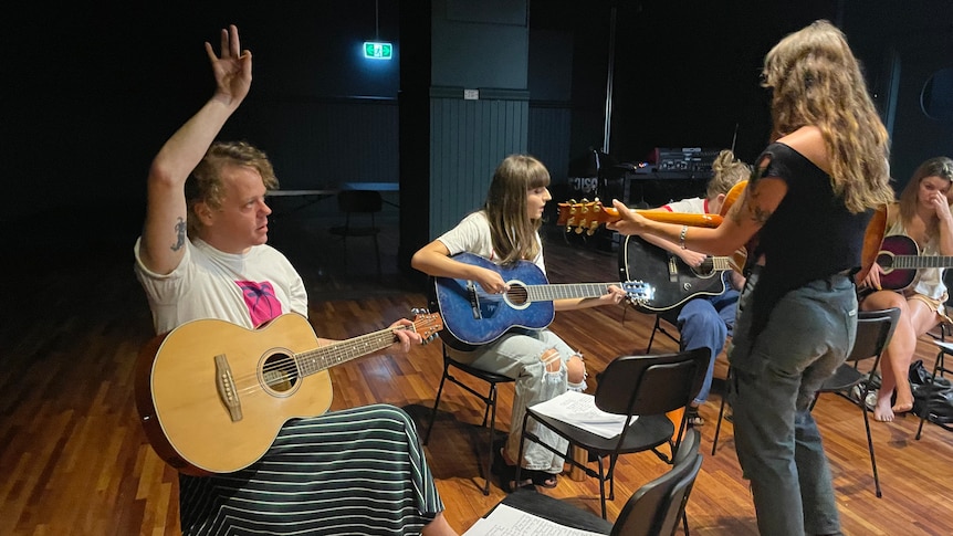 A woman with her back to the camera plays guitar in front of two people sitting down also holding guitars.