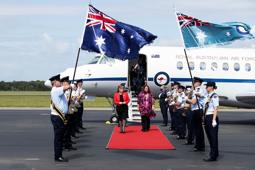 Female ministers Reynolds and Price are welcomed by red carpet and guard of honour at airport.