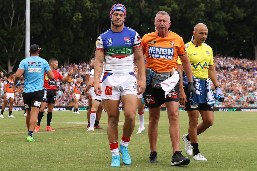 A rugby league player wearing headgear is escorted off the field by support staff.
