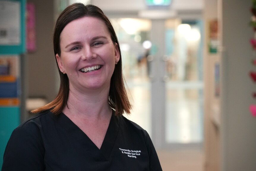 A nurse in navy scrubs smiling in the corridor of a hospital ward