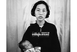 A black and white image of a Cambodian woman holding a baby with Khmer script and a number around her neck.