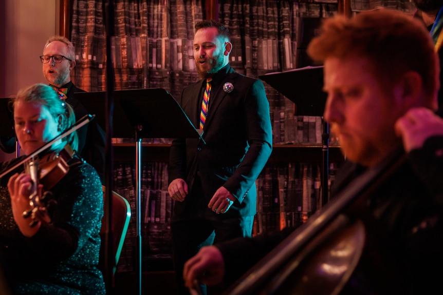 Four members of the ensemble wear all black with rainbow accessories, performing in a dimly lit room.