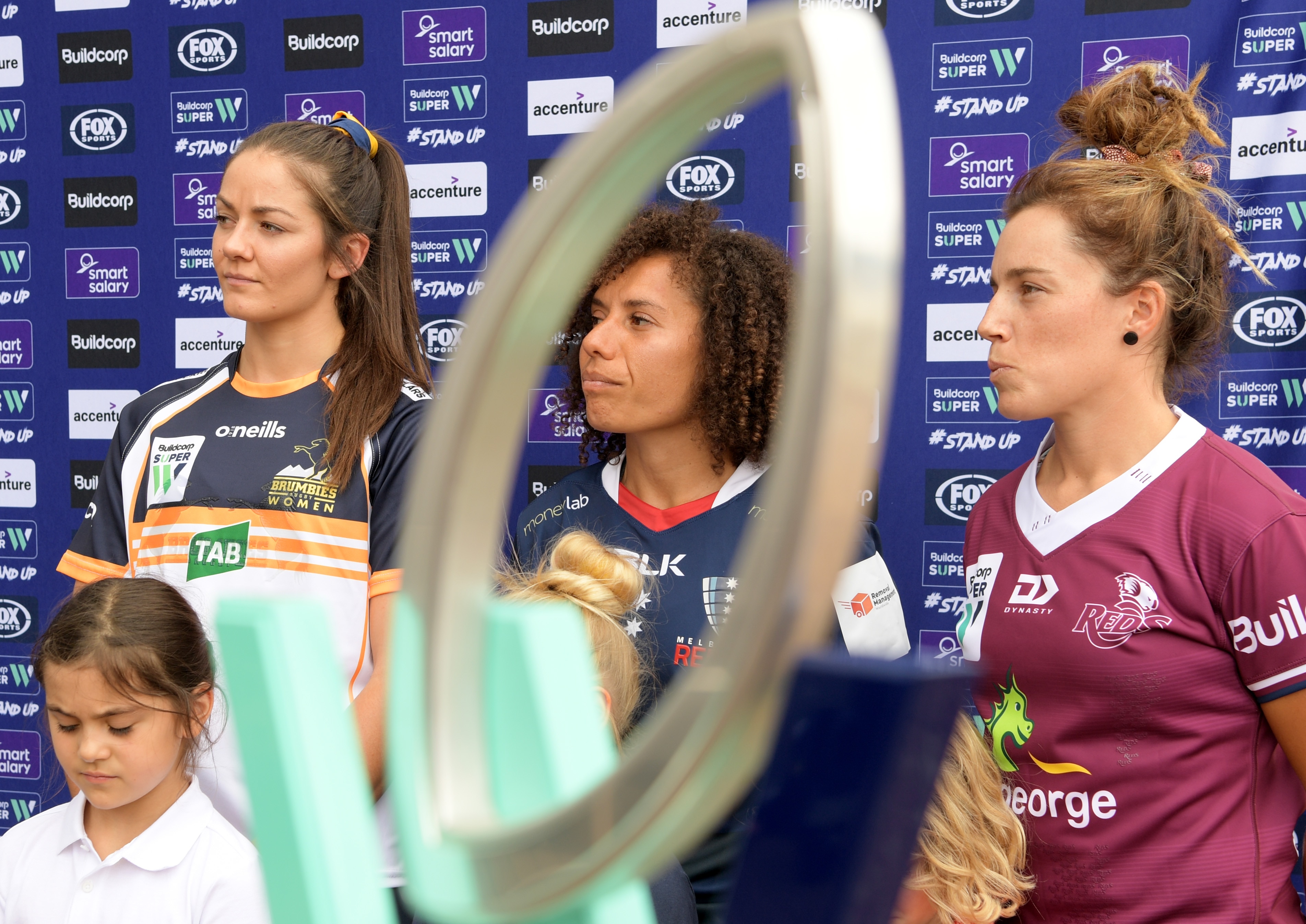Three women pose in front of a Super W trophy with young girls in front of a banner.