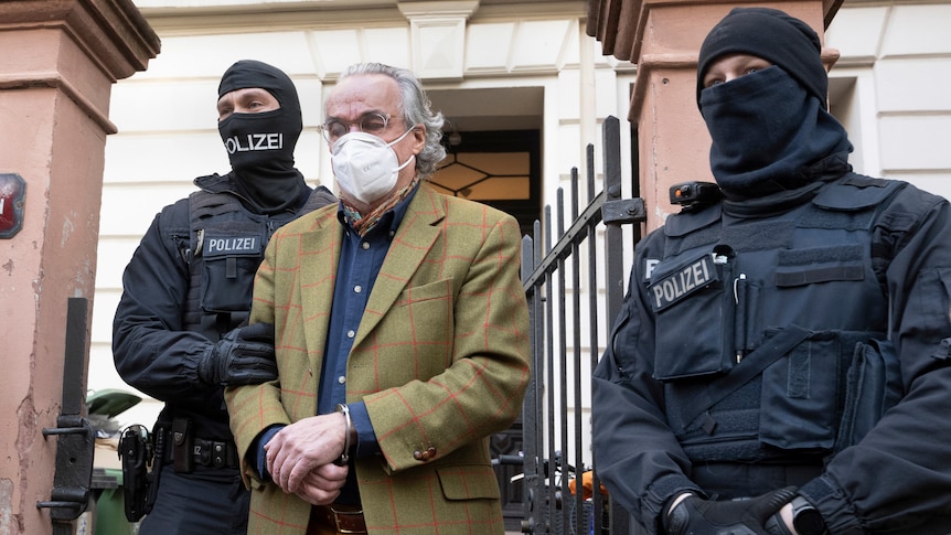 Two armed police in tactical gear and balaclavas walk a handcuffed man in a mask and tweed jacket out o a home. 