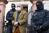 Two armed police in tactical gear and balaclavas walk a handcuffed man in a mask and tweed jacket out o a home. 