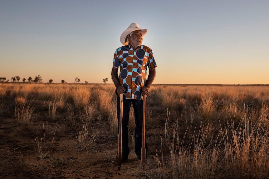 A photo of Indigenous artist Ngarralja Tommy May standing in remote country as the sun sets.