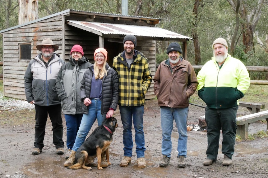 Four men and two women standing in front of a timber hut with iron roof, and a dog at their feet.