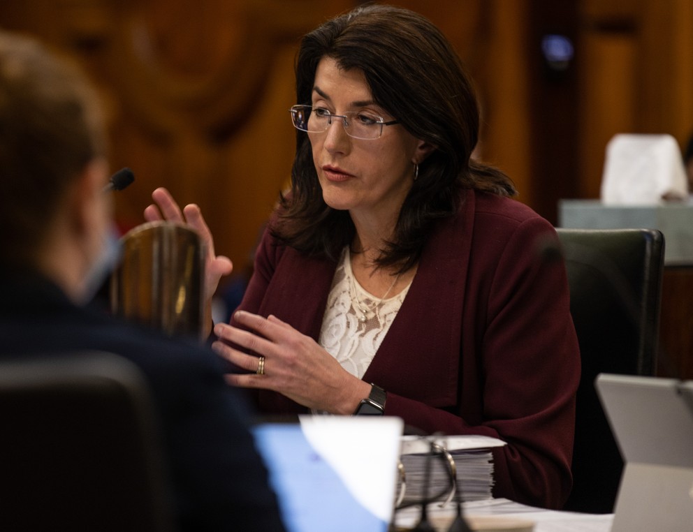 A woman with dark hair sits at a desk and speaks