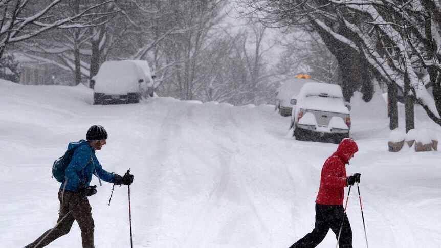 Two people in snow gear, with walking poles, trudge across a road covered in snow. The cars behind them are all but buried.