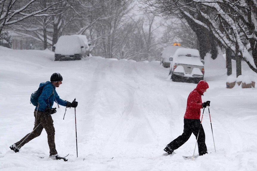 Two people in snow gear, with walking poles, trudge across a road covered in snow. The cars behind them are all but buried.