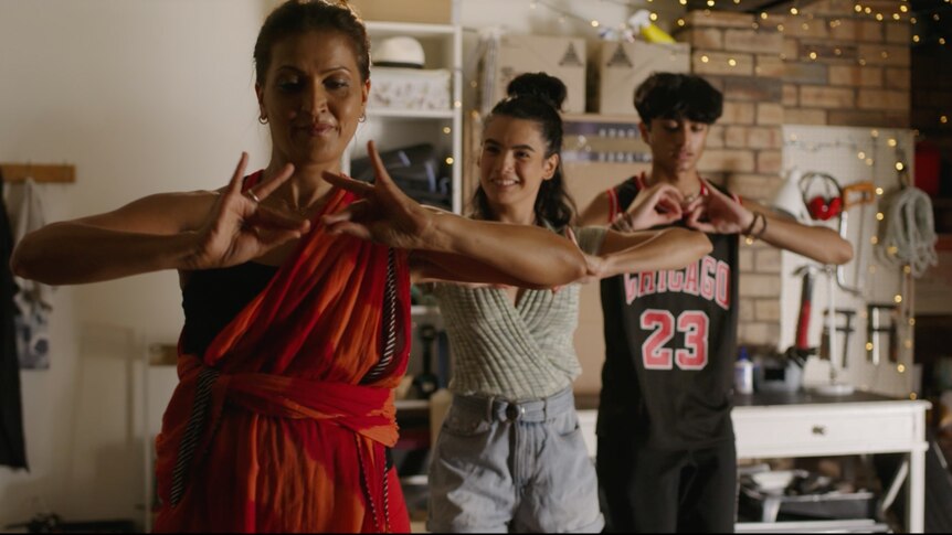 Three figures - a woman in a red sari and two teens - dance in front of a tool area in someone's home.